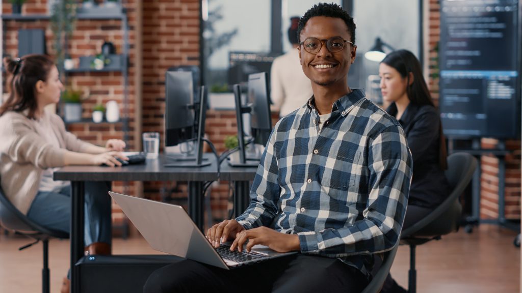 Portrait of artificial intelligence app developer sitting down typing on laptop fixing glasses looking up and smiling at camera. Programer using portable computer innovating cloud computing.