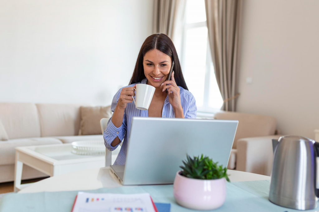 Self employed woman working with her phone and laptop at home with a cup of coffee Happy woman using mobile phone while working at home with laptop Smiling woman messaging with smartphone